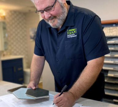 A bearded man wearing a blue "Fred" polo shirt smiles while making notes on architectural diagrams. He holds different types of tiles in one hand and a pen in the other.